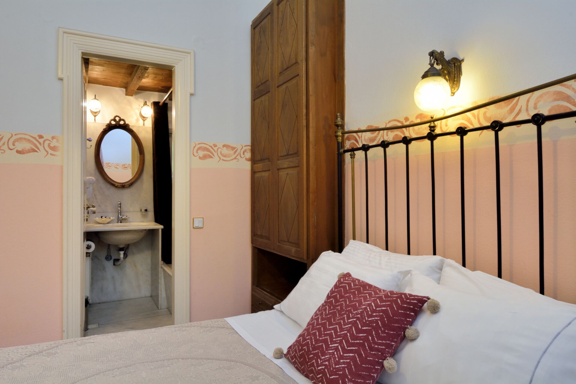 A bedroom with a bed, a nightstand, a mirror, a wooden wardrobe, and a wall-mounted light fixture.