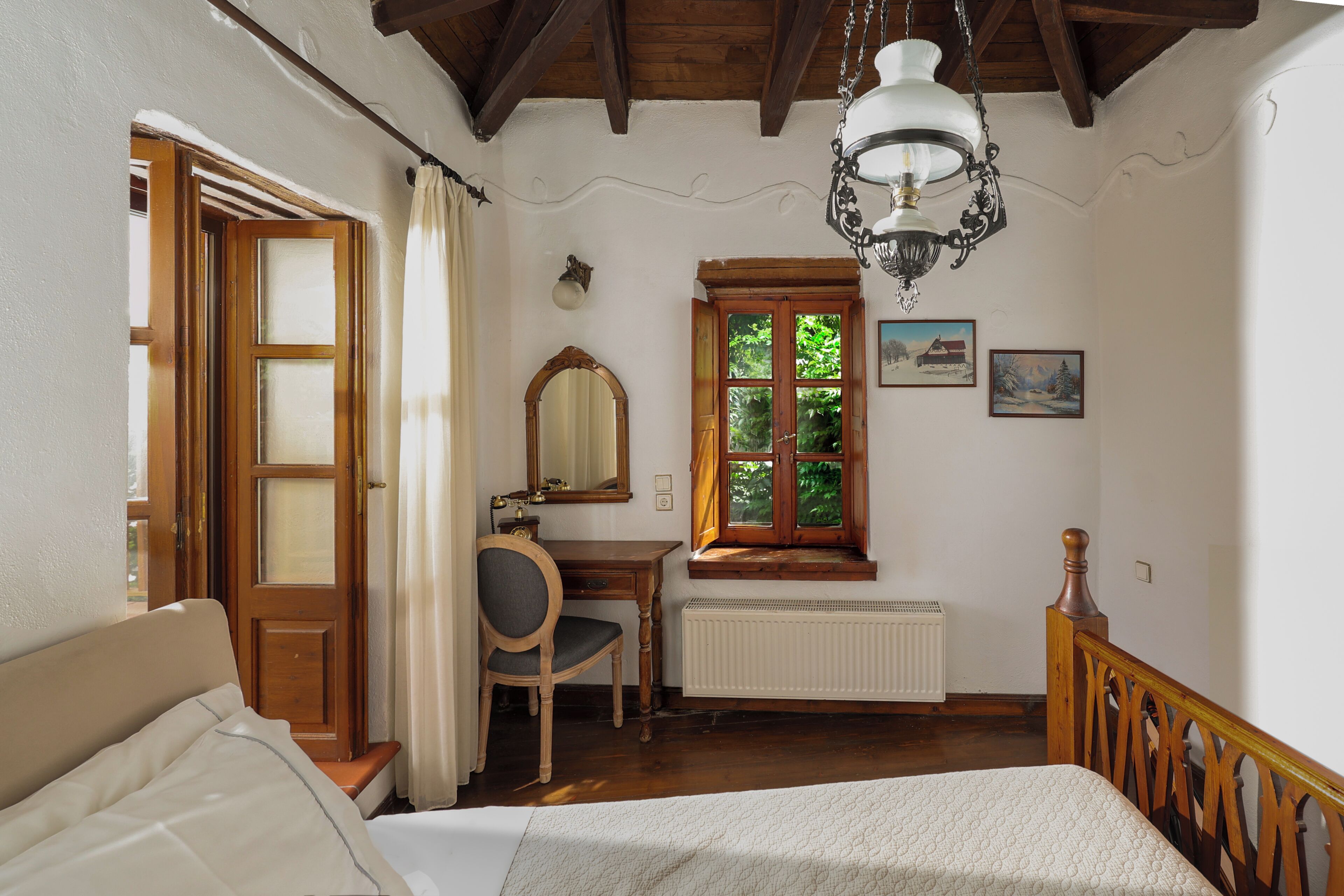 A bedroom with a bed, a desk, a chair, a window with curtains, a radiator, and a wooden ceiling.