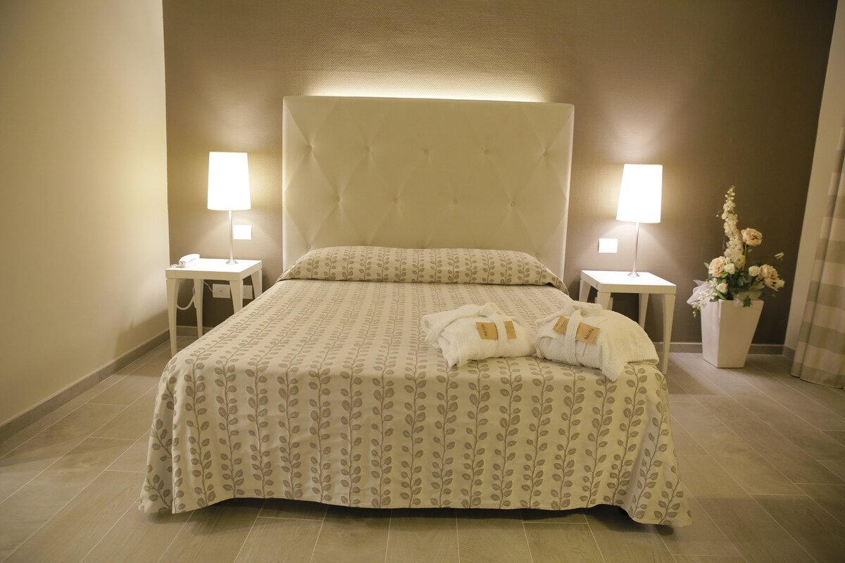 A neatly made bed with a patterned bedspread, two bedside lamps, and a vase with flowers on the right.