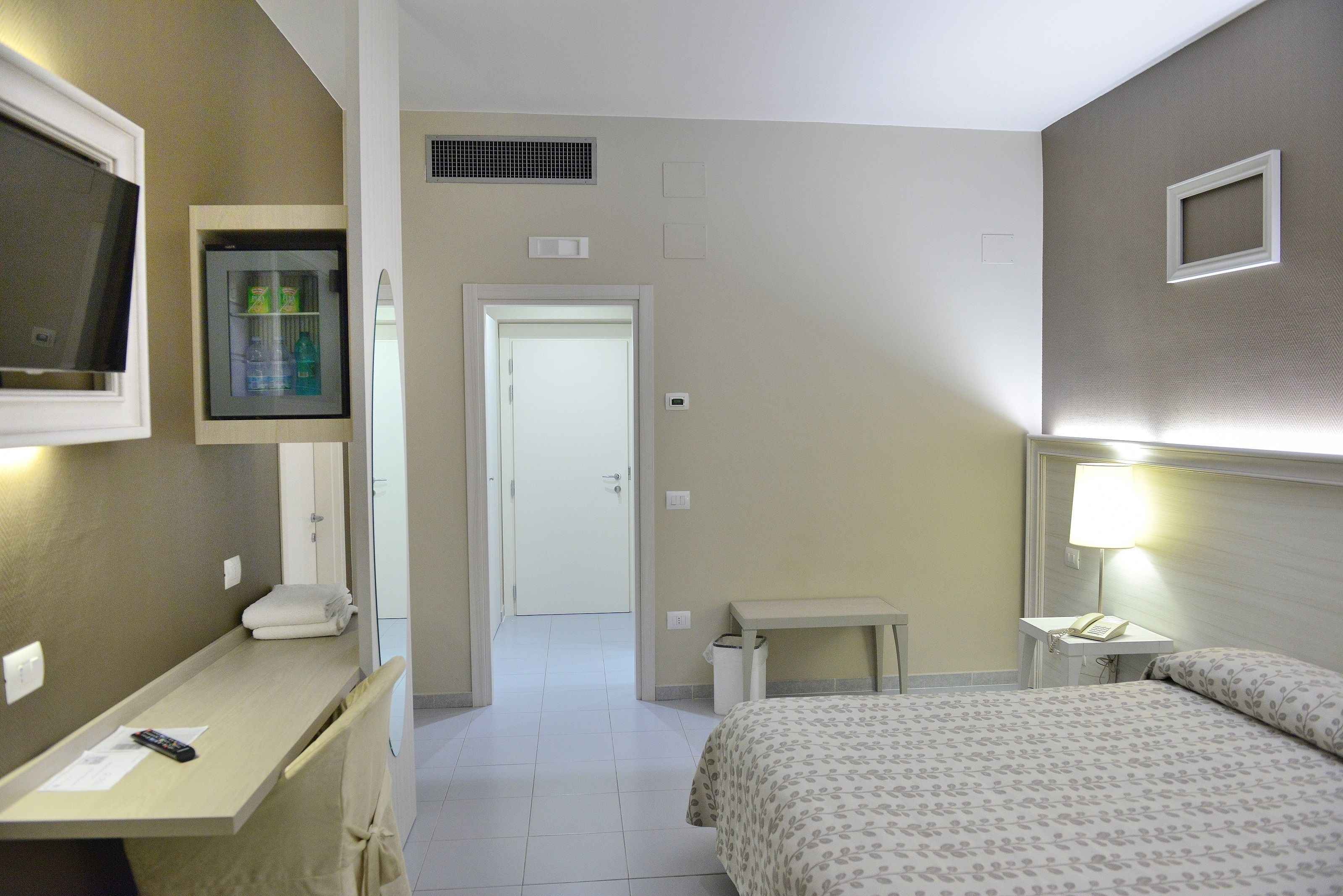 Habitación de hotel con cama, mesita de noche, un pequeño escritorio con silla, televisor en la pared y una puerta que da a otra habitación.