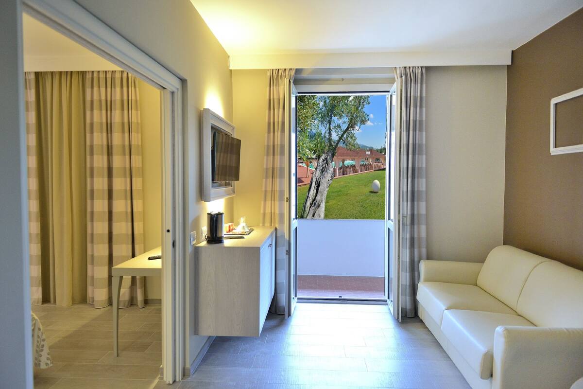 A hotel room with a sofa, a TV, a desk, and a view of a garden through a sliding door.