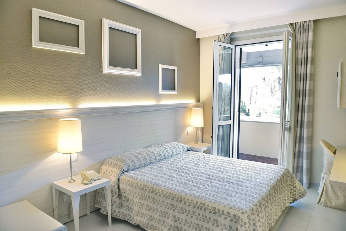 A hotel room with a bed, two bedside lamps, a nightstand, a window with curtains, and a door.