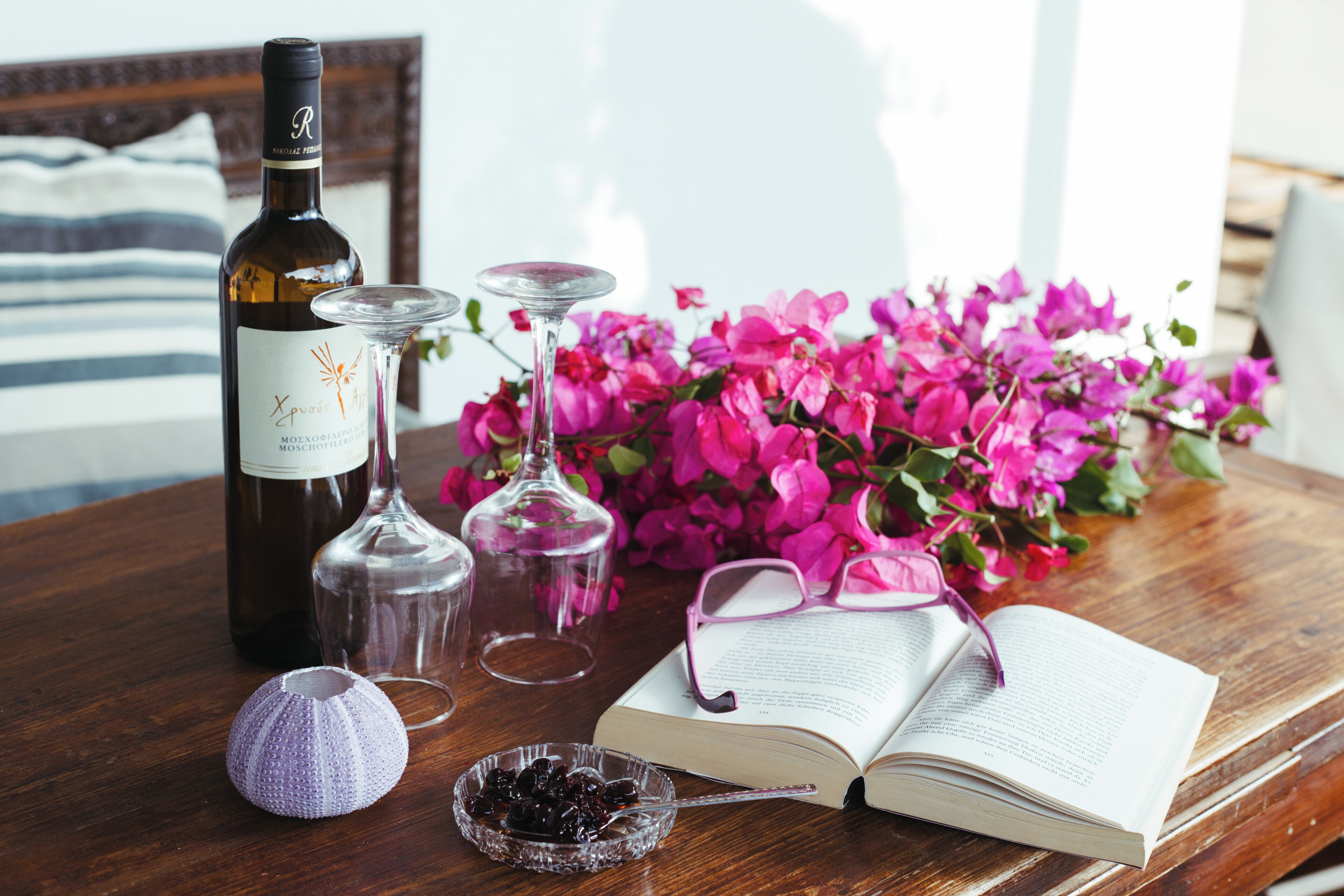 A wooden table with a wine bottle, two wine glasses, a book, a pair of glasses, and a bowl of berries.