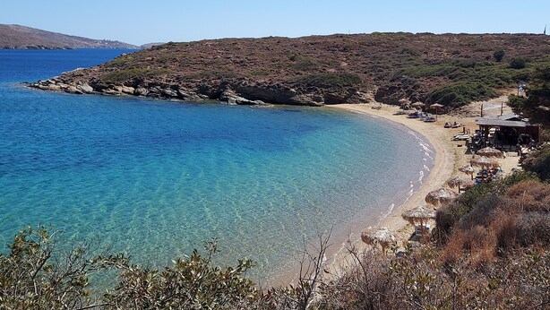 A coastal beach with clear blue waters, rocky terrain, and a small beachside structure with umbrellas. - Villa Arni (Andros)