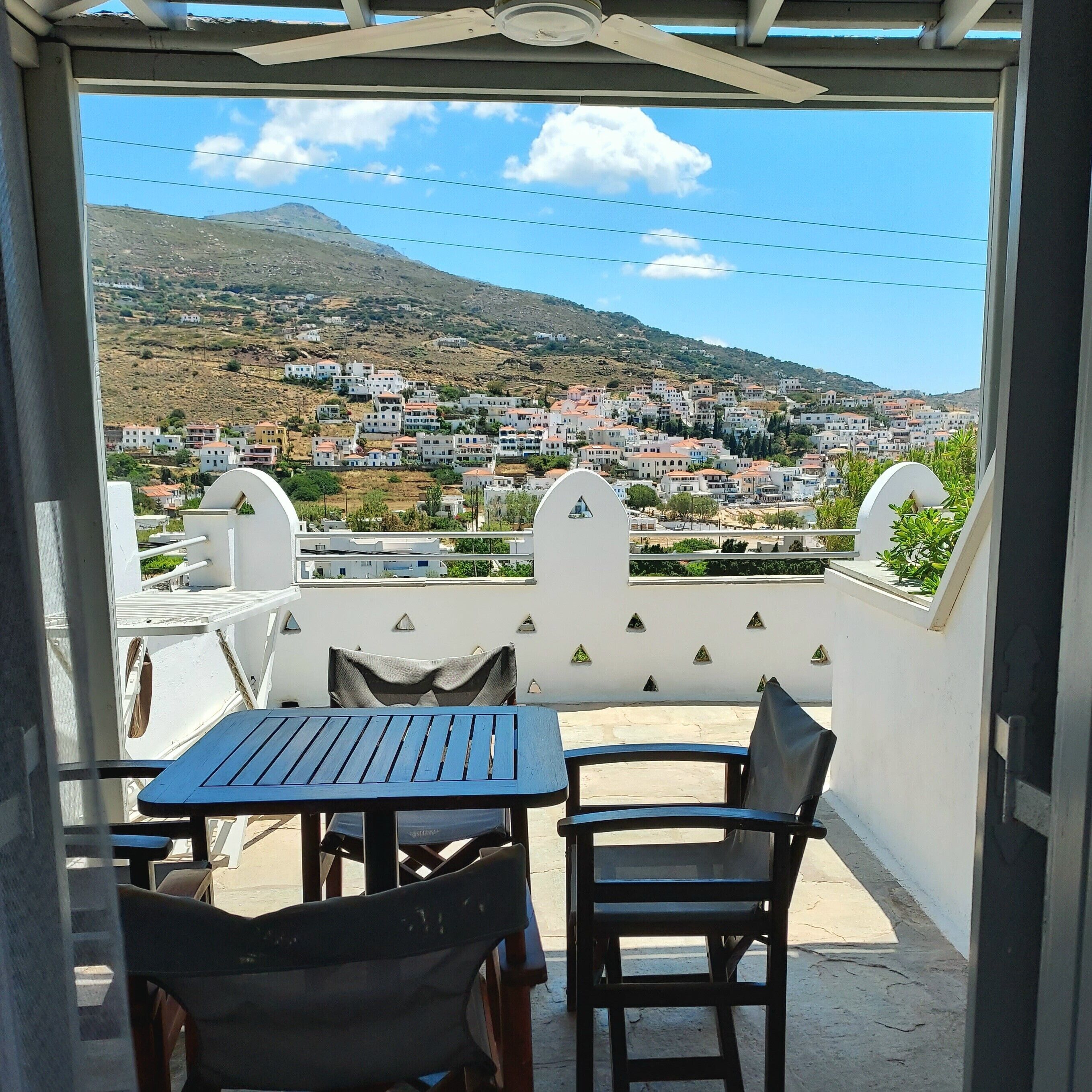 A balcony with a table and chairs, overlooking a hillside town with buildings and a mountain in the distance.