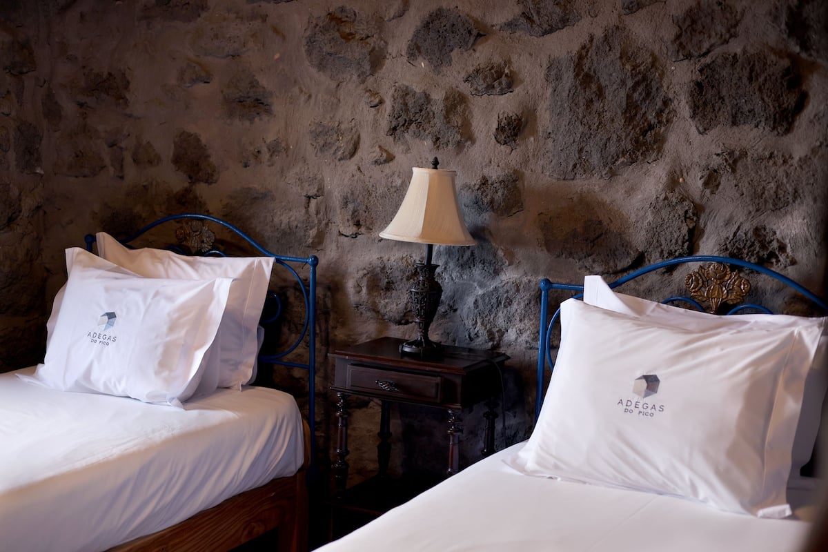 A room with two beds, each with white bedding and pillows, a stone wall, a nightstand with a lamp, and a small table.