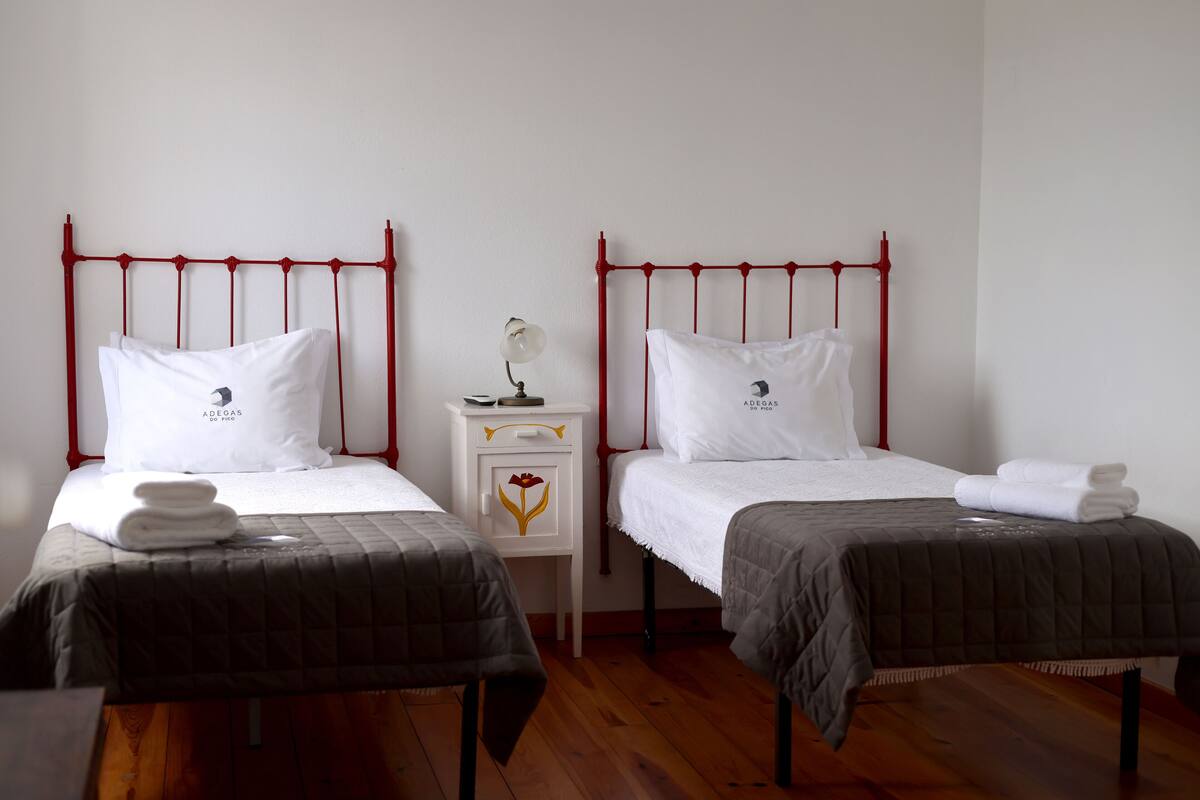 A hotel room with two beds, each with a red metal headboard, white bedding, and a small bedside table with a lamp.