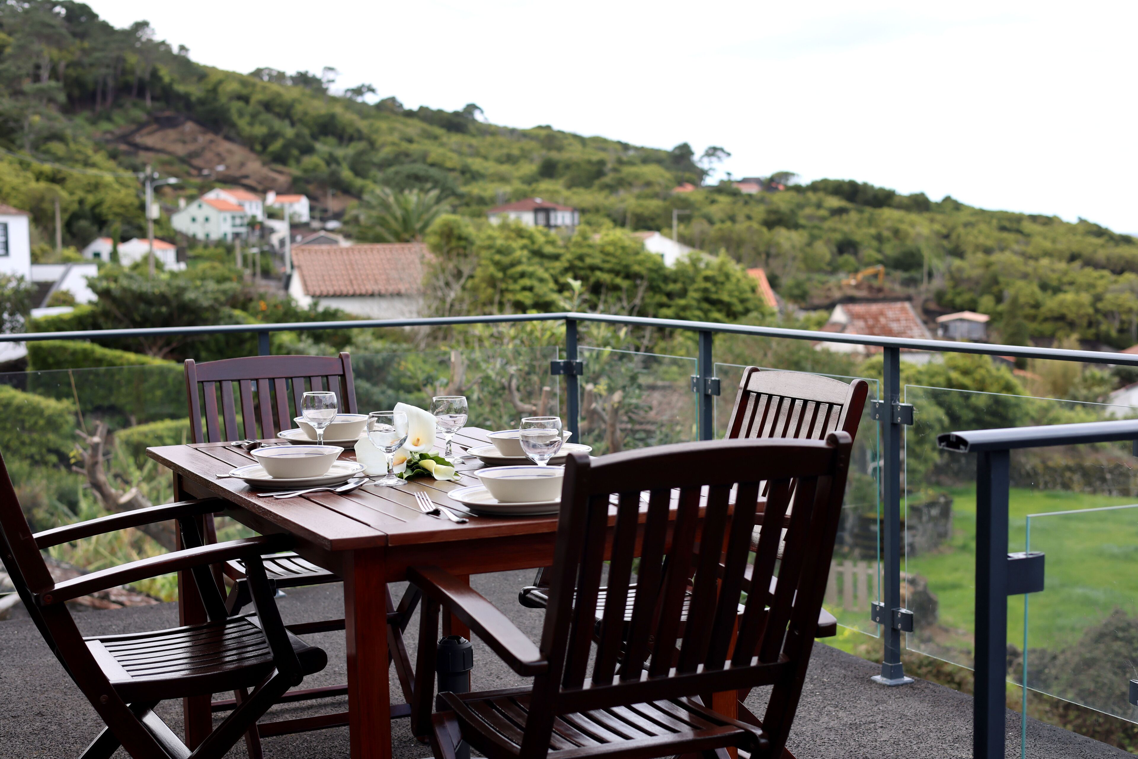 A dining table set with plates, glasses, and cutlery, overlooking a lush green landscape with houses in the background.