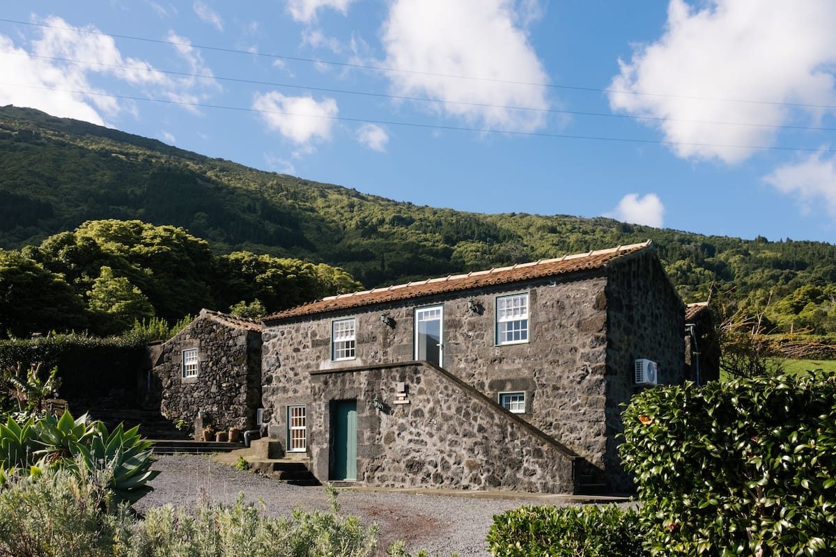 A stone building with a green door and windows, surrounded by greenery and hills.