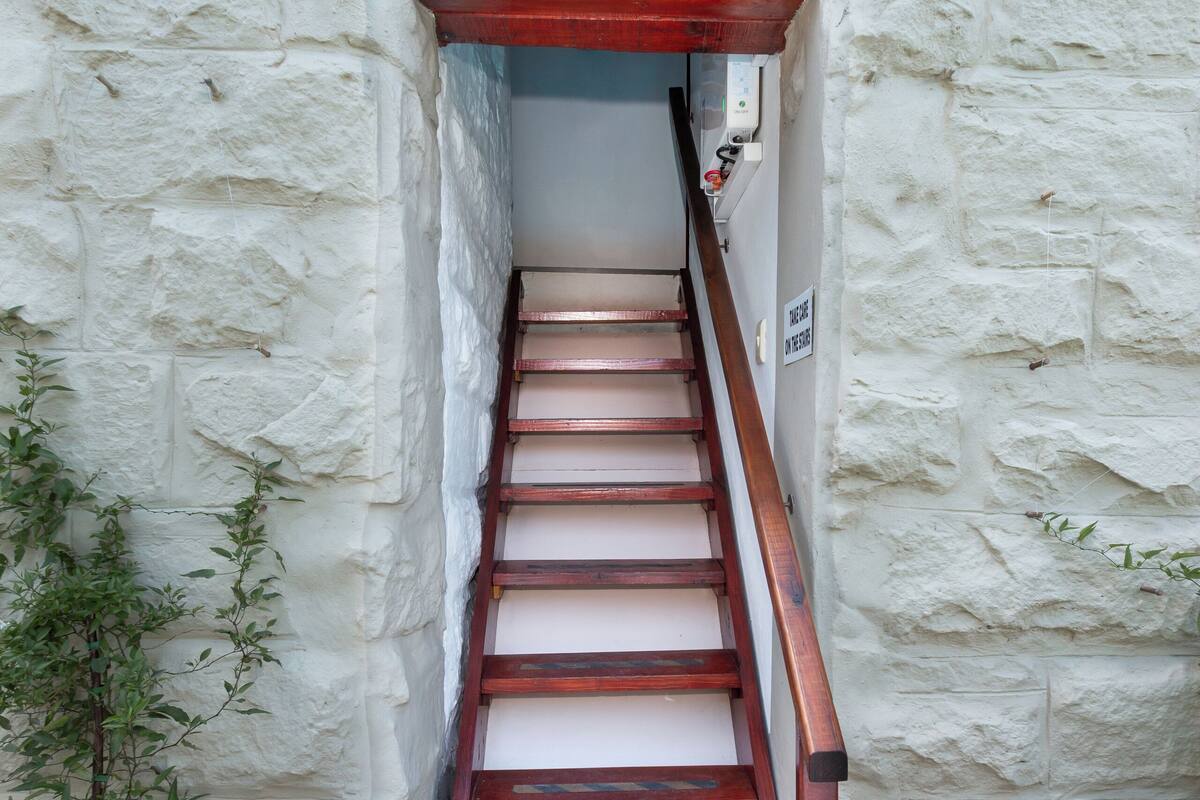 Uma escadaria estreita com corrimãos e degraus de madeira, que leva a uma porta com uma placa em cima.