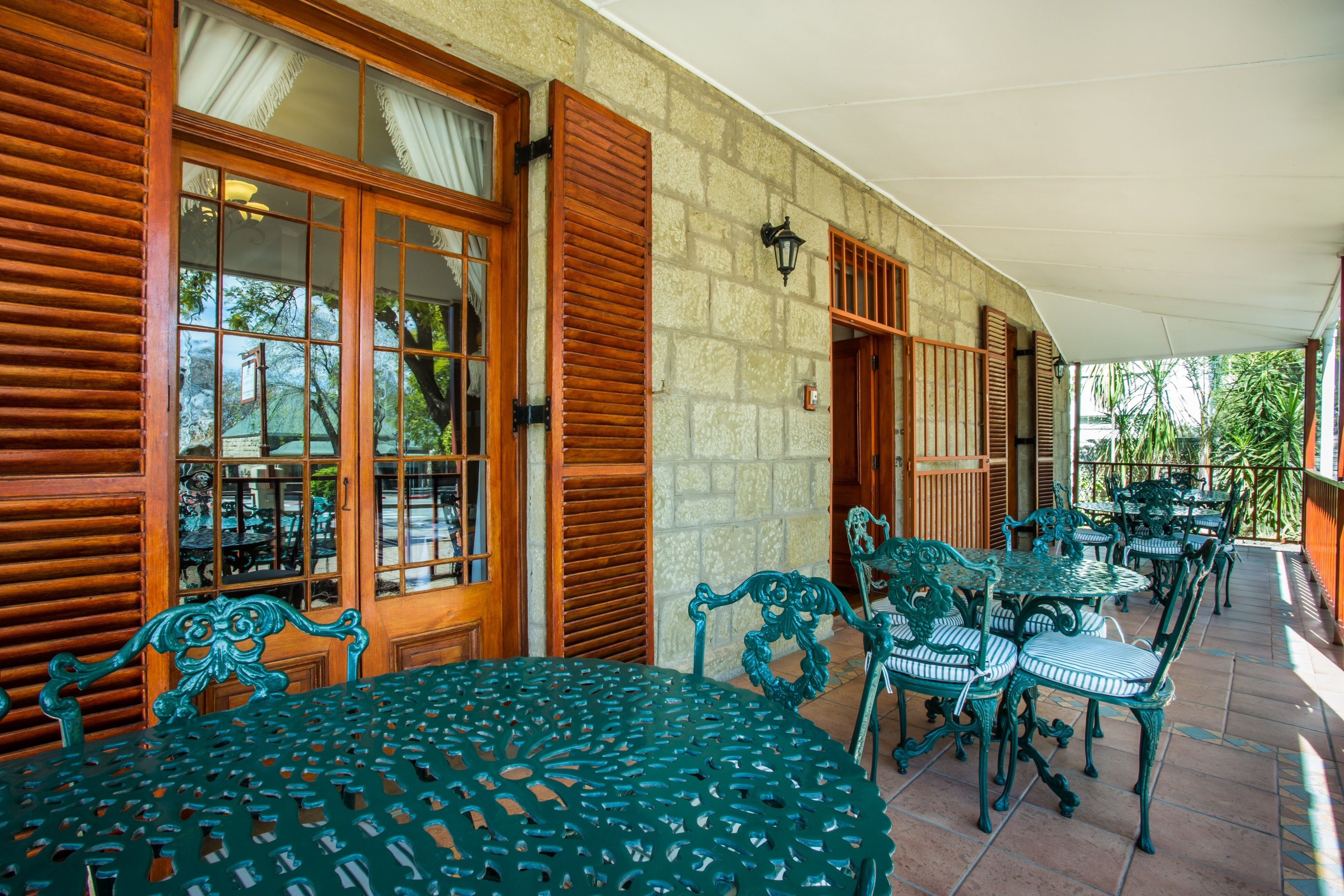 Un patio cubierto con contraventanas de madera, mesas y sillas de metal verde y una pared de piedra.