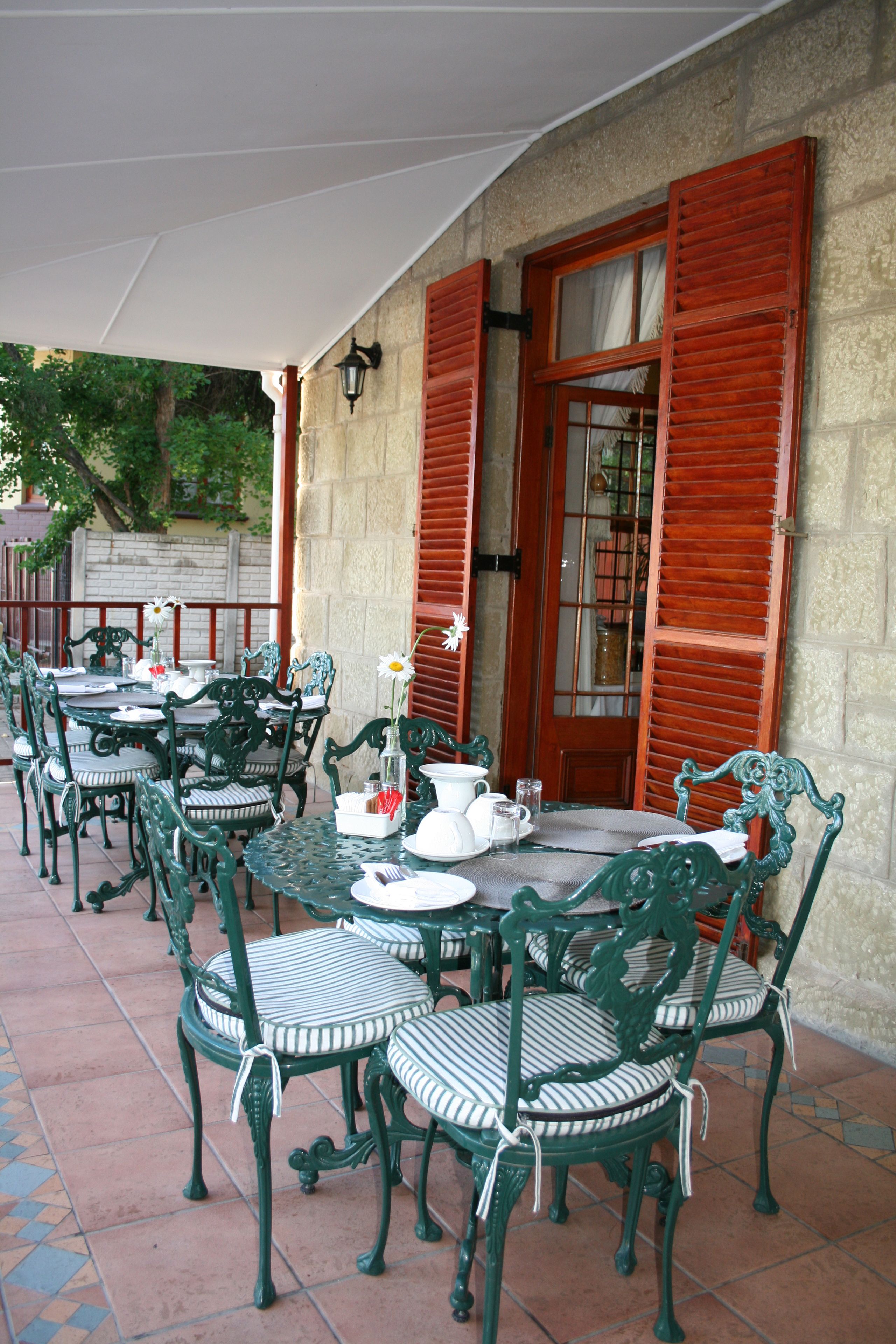 Une terrasse extérieure avec plusieurs tables et chaises, un parasol blanc et un mur de pierre agrémenté de volets rouges.