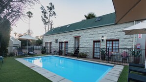 A swimming pool with a stone building in the background, surrounded by a fenced area with lounge chairs.
