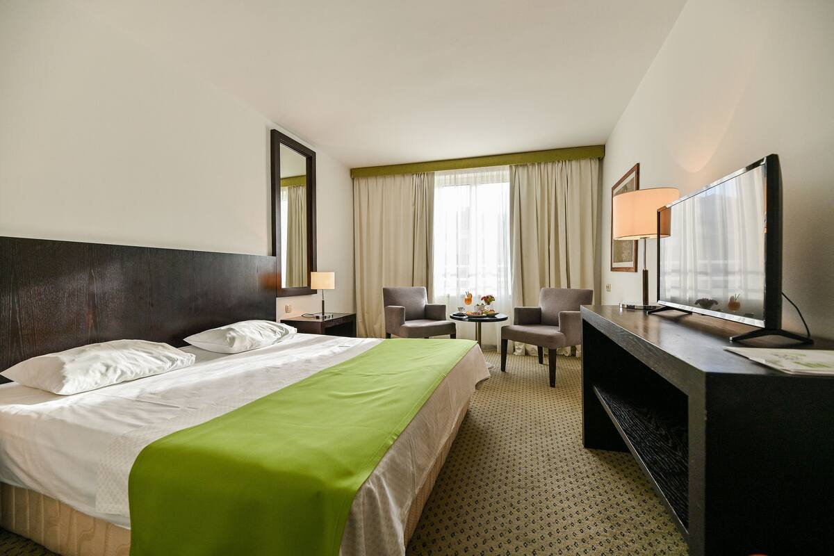 Une chambre d’hôtel comprenant un lit, deux fauteuils, un bureau avec une télévision et une petite table avec une lampe.