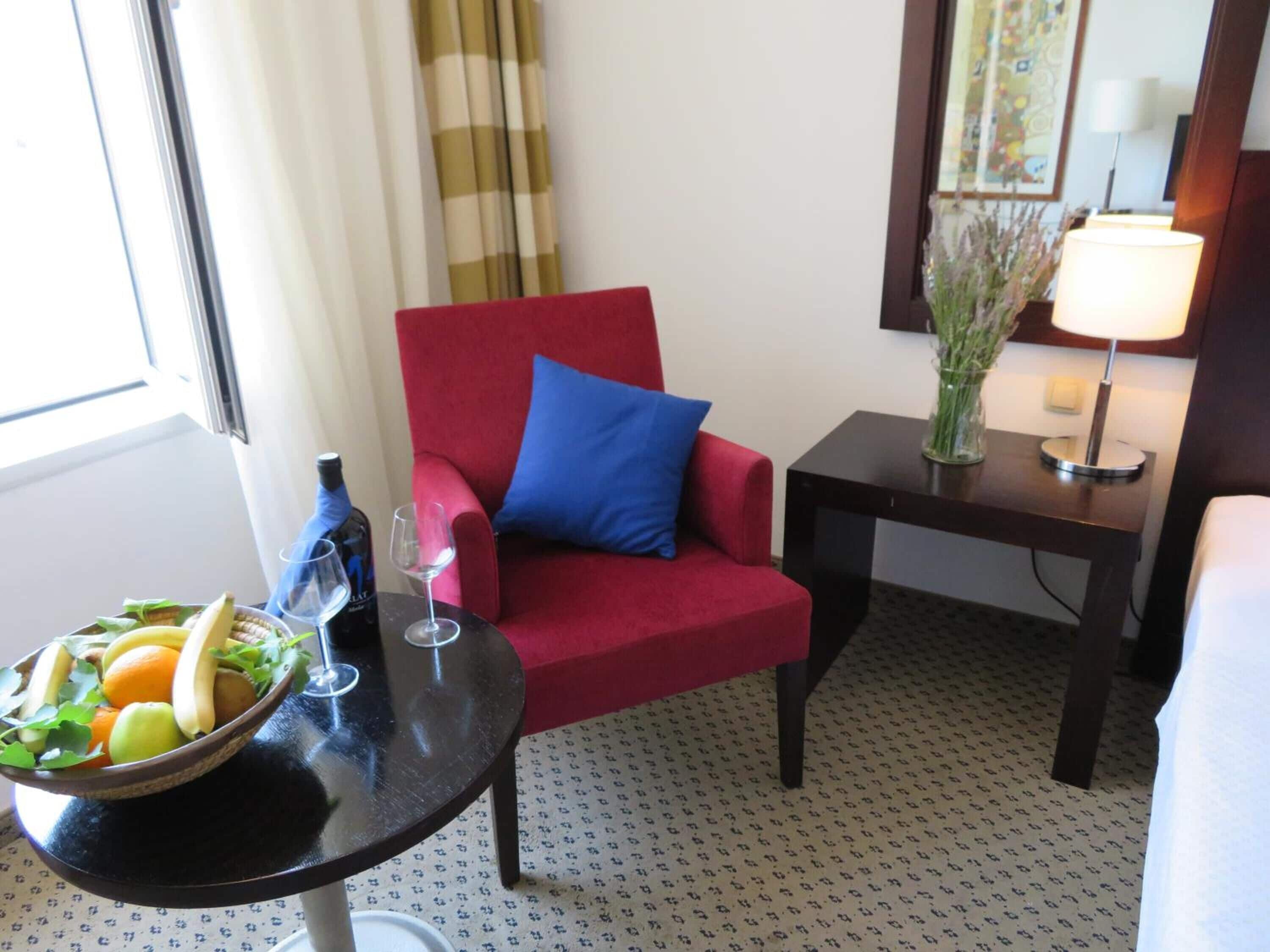 A hotel room with a red armchair, a small table with a bowl of fruit and wine glasses, a lamp, and a mirror.