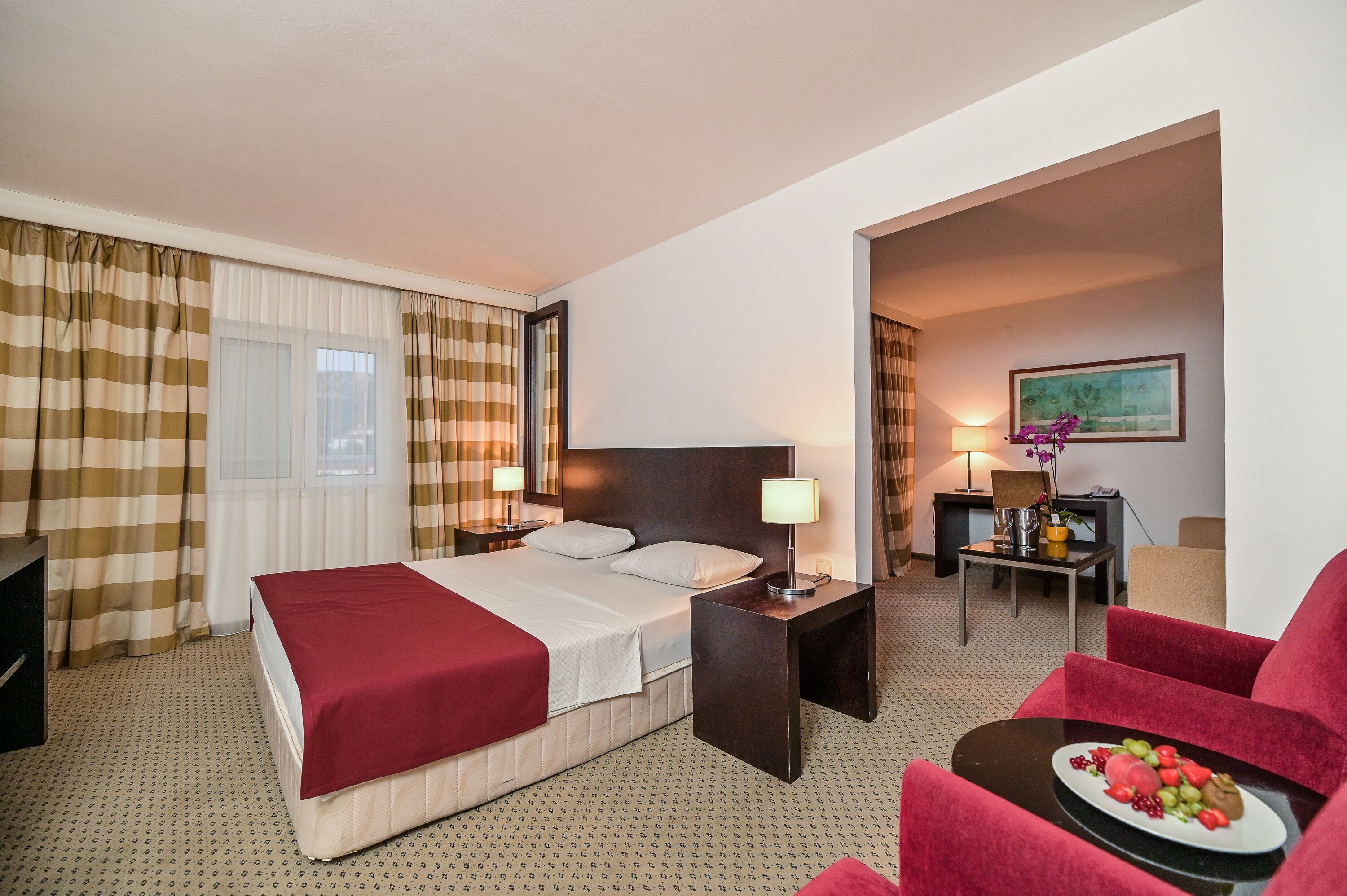 A hotel room with a bed, a desk, a chair, a lamp, and a dining table with fruit.