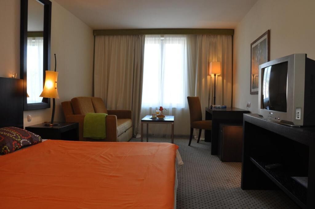 A hotel room with a bed, two armchairs, a small table, a desk, a television, and a window with curtains.