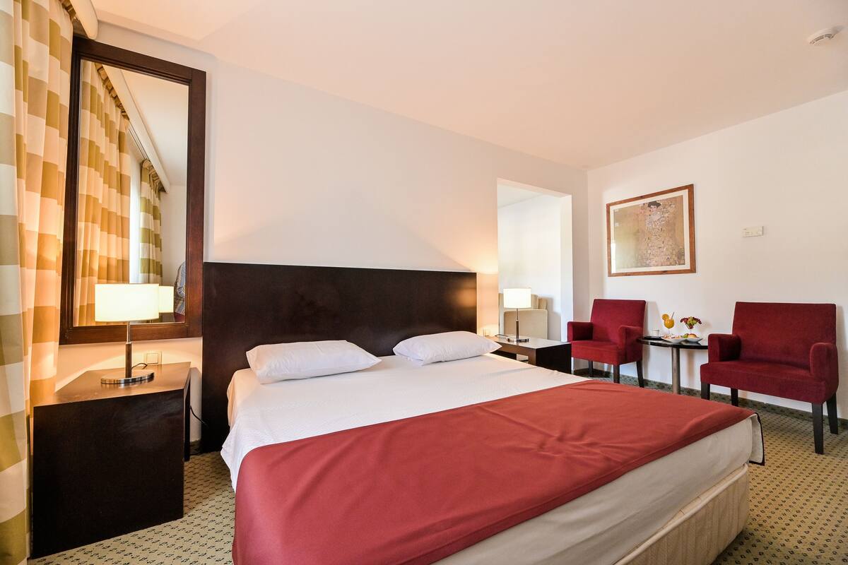 Une chambre d’hôtel avec un grand lit, deux tables de chevet avec des lampes, un fauteuil rouge, une petite table avec un vase et un tableau encadré au mur.