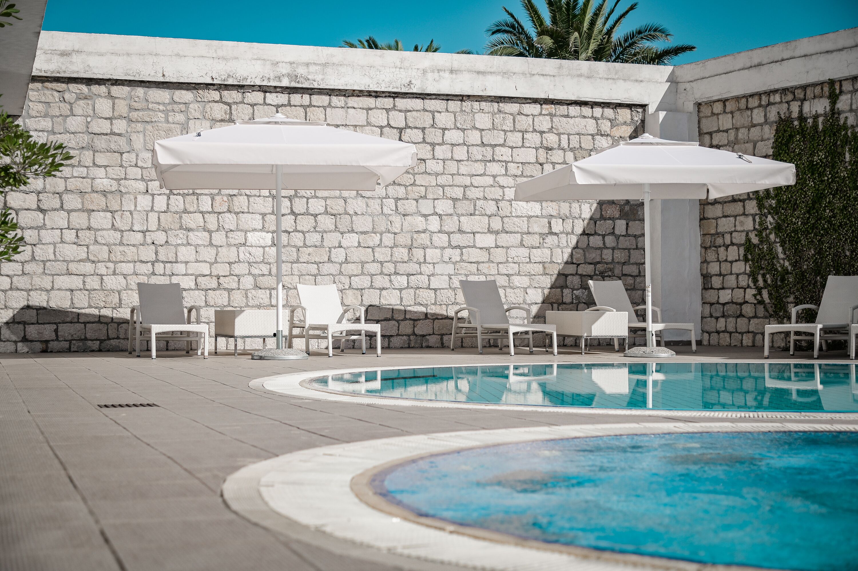 Un espace piscine agrémenté de chaises longues blanches et de parasols, le tout entouré d’un mur de pierre.