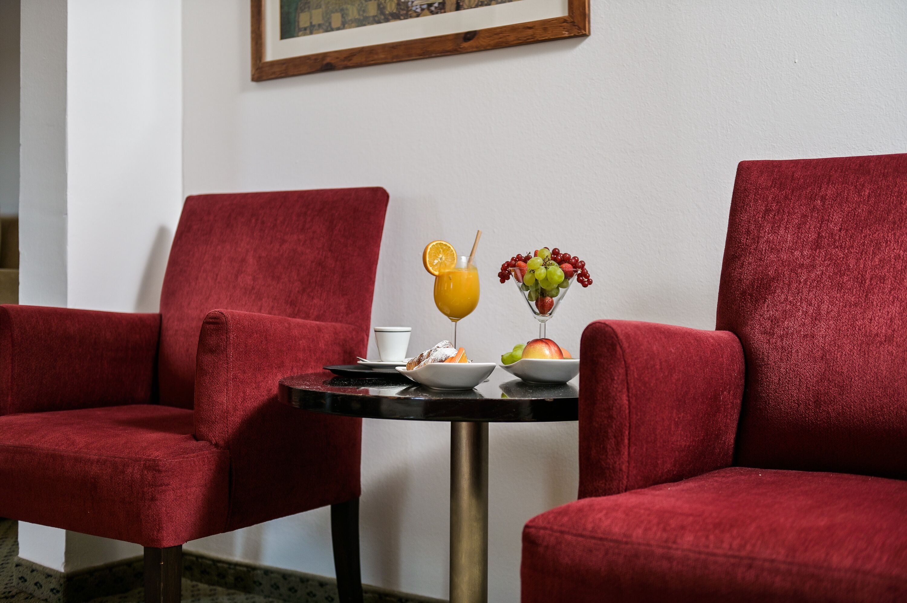 Ein Zimmer mit zwei roten Polstersesseln, einem kleinen runden Tisch, einer weißen Tasse, einem Glas Orangensaft, einer Obstschale und einer Vase mit mehr Früchten.