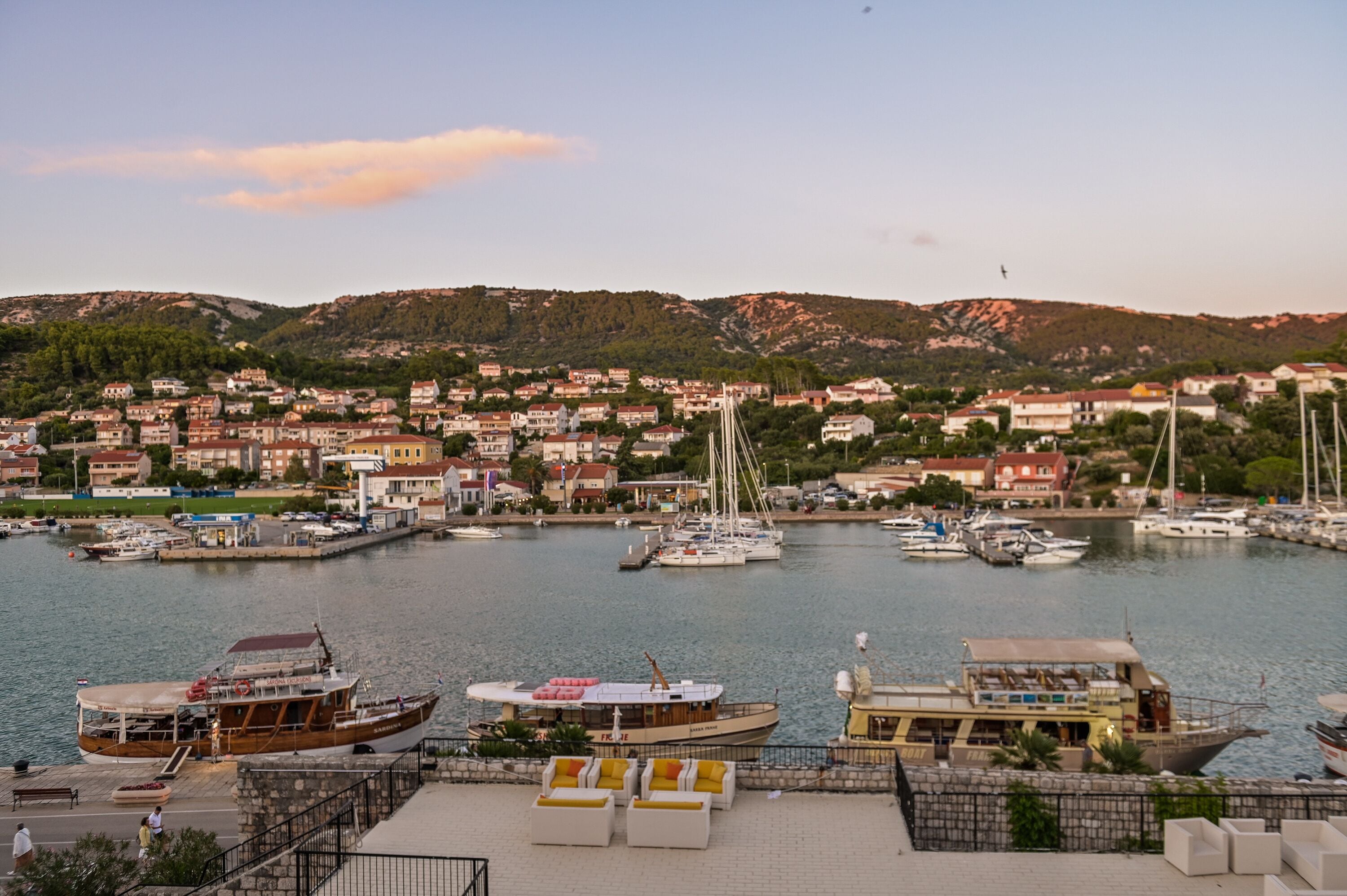 A marina with boats docked, a waterfront promenade with seating, and a backdrop of residential buildings and hills.