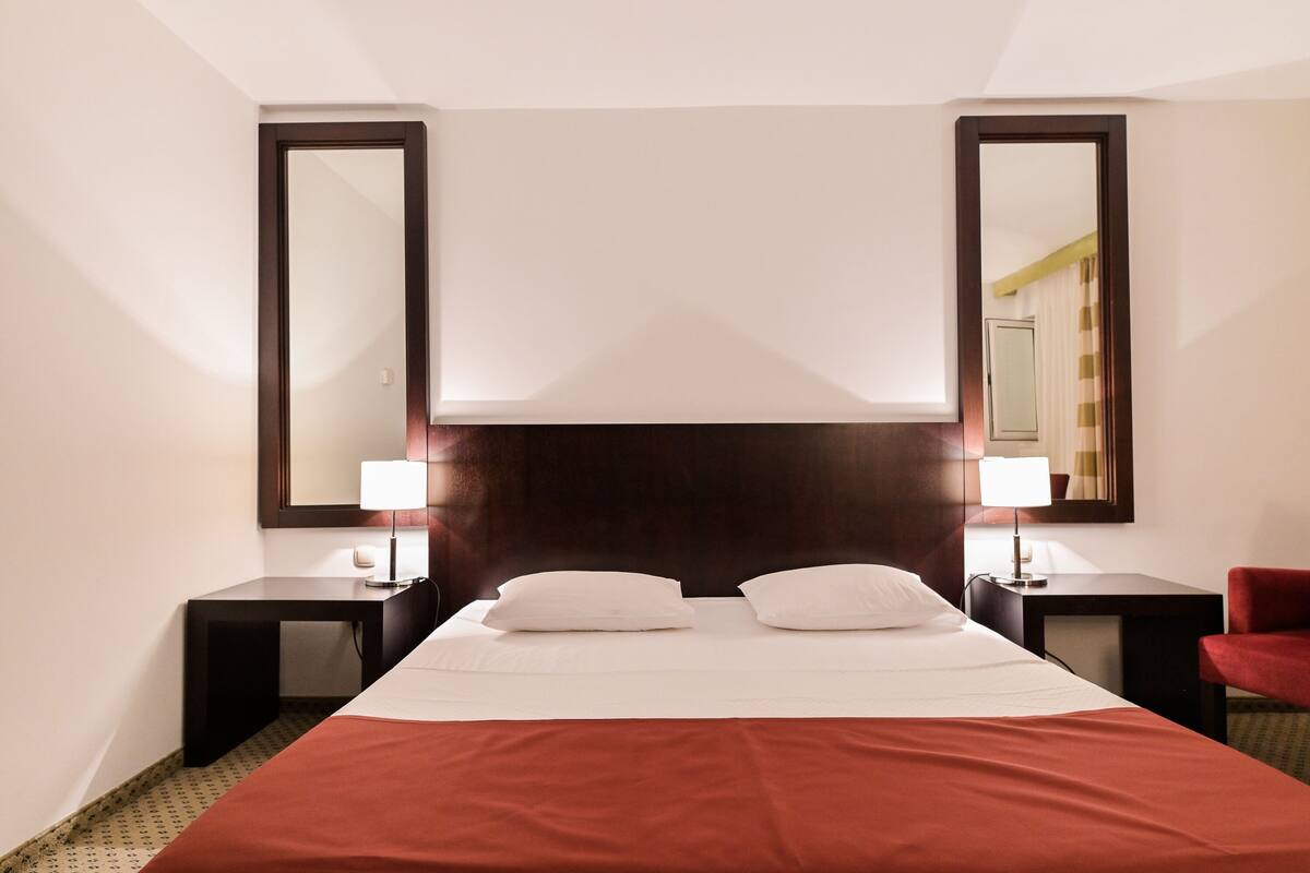 Une chambre d’hôtel avec un grand lit, deux tables de chevet avec des lampes, un fauteuil rouge et deux grands miroirs sur les murs.