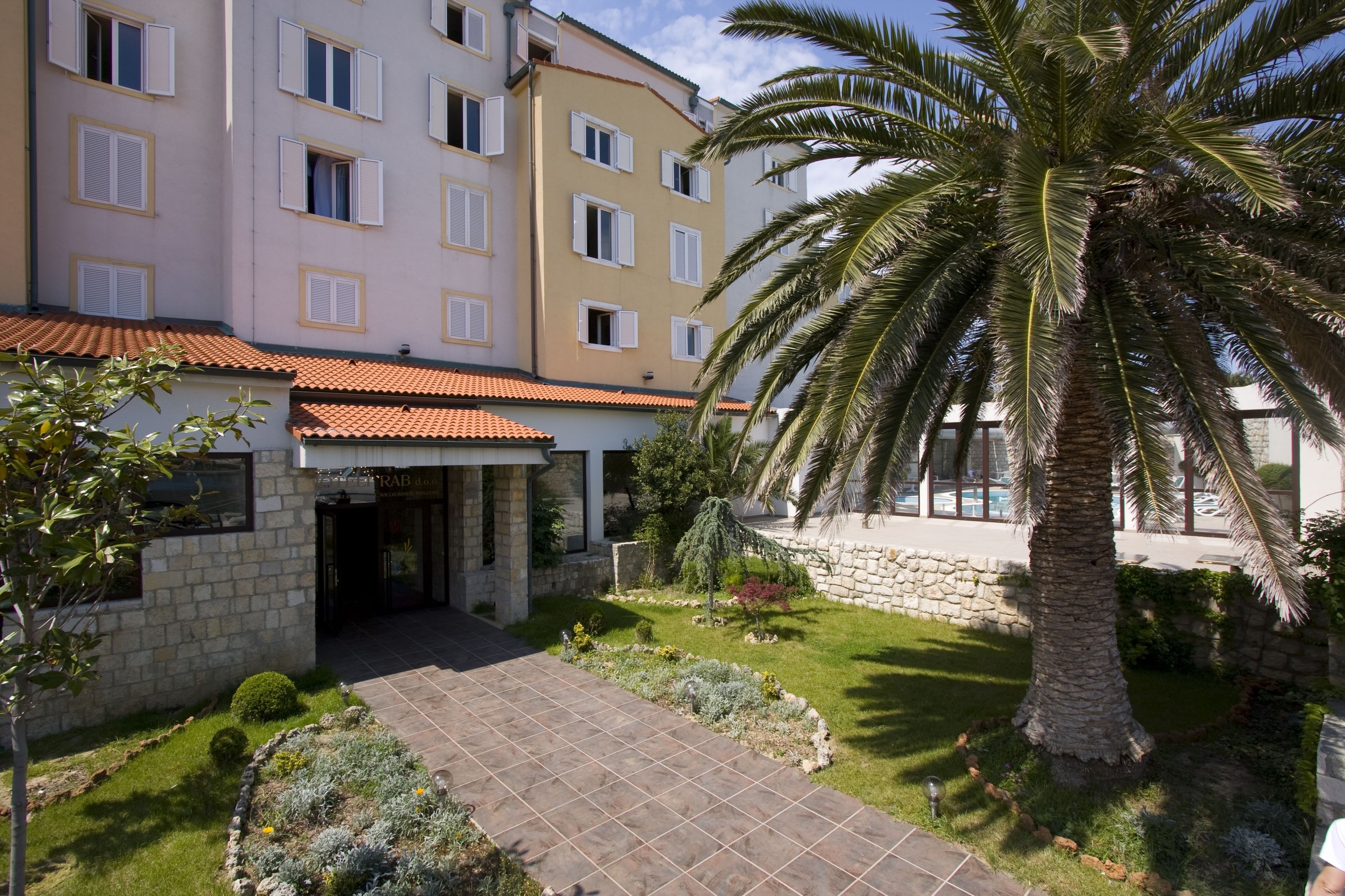 Un hotel de varios pisos con fachada de piedra, techo de tejas y un jardín bien cuidado con palmeras y arbustos.