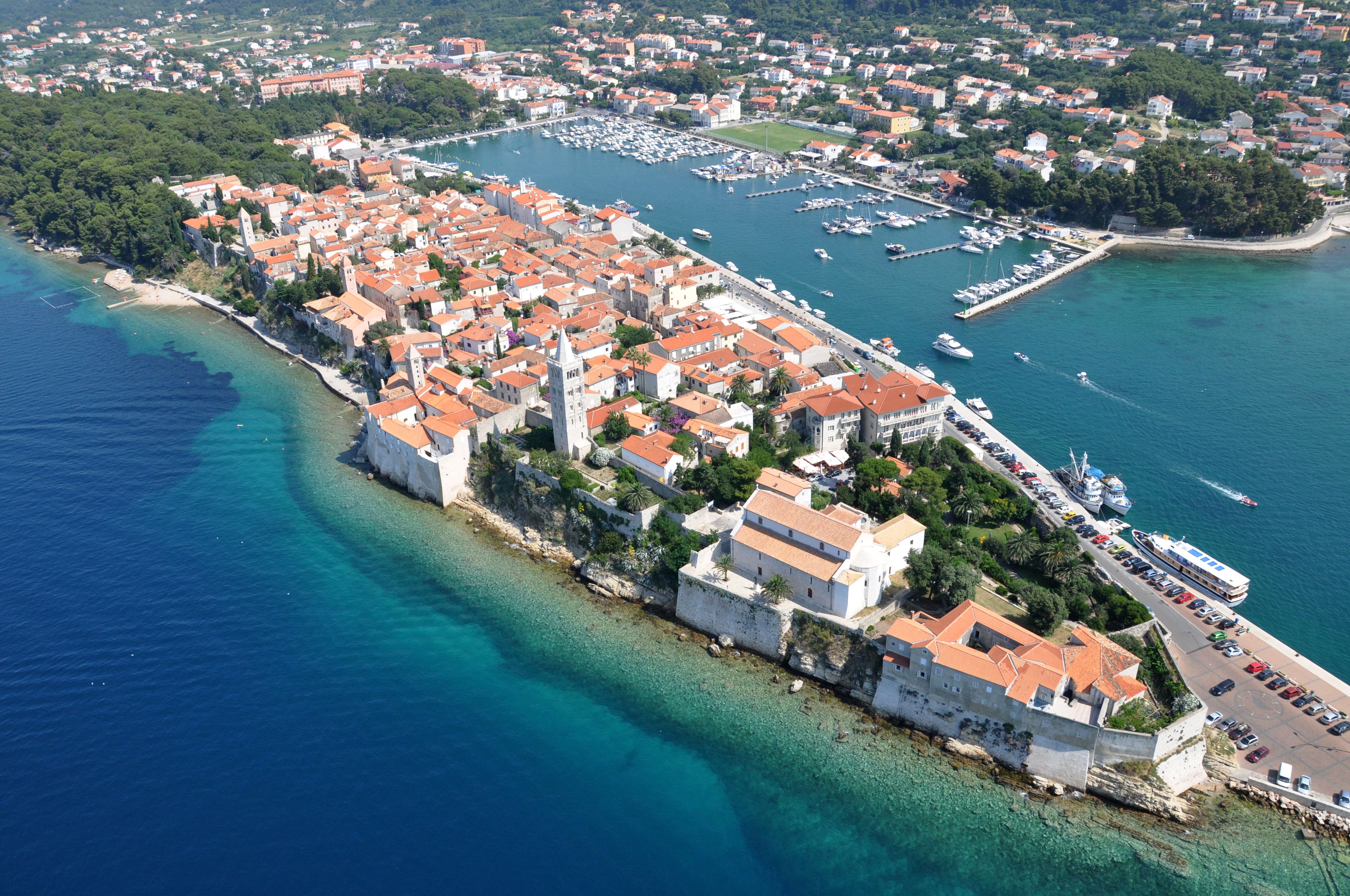An aerial view of a coastal town with historic buildings, a marina, and a clear blue sea.
