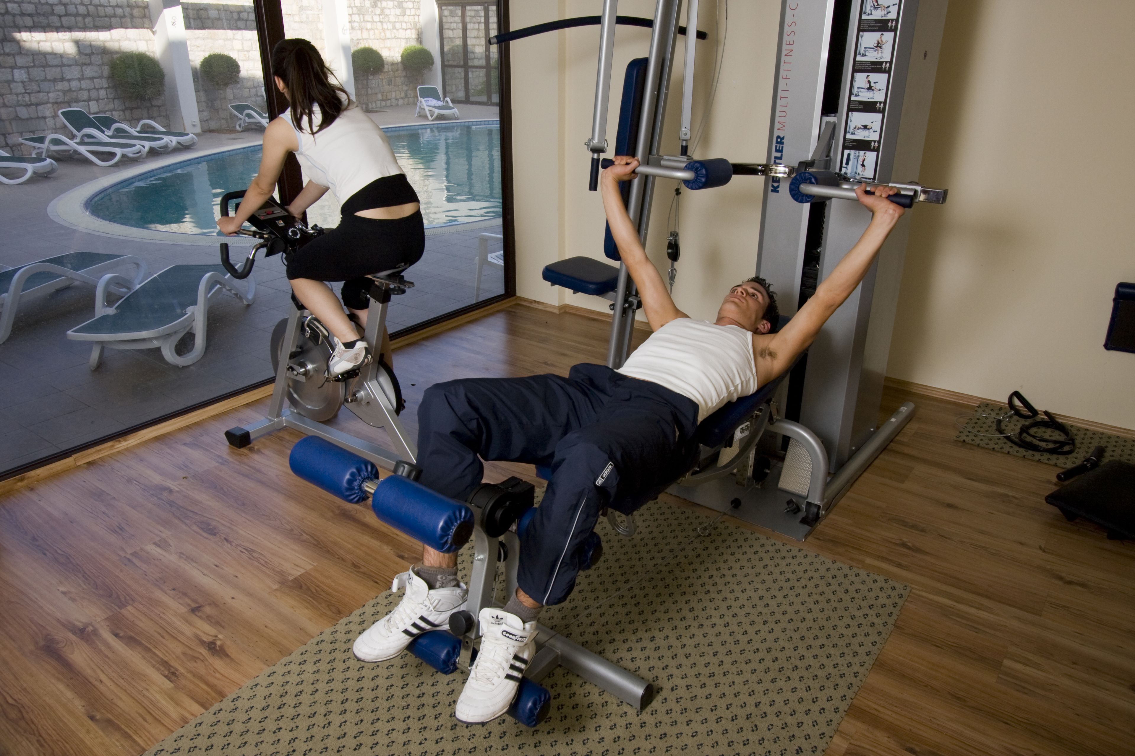 Ein Fitnessstudio mit Blick auf einen Pool, Trainingsgeräten und Holzboden.