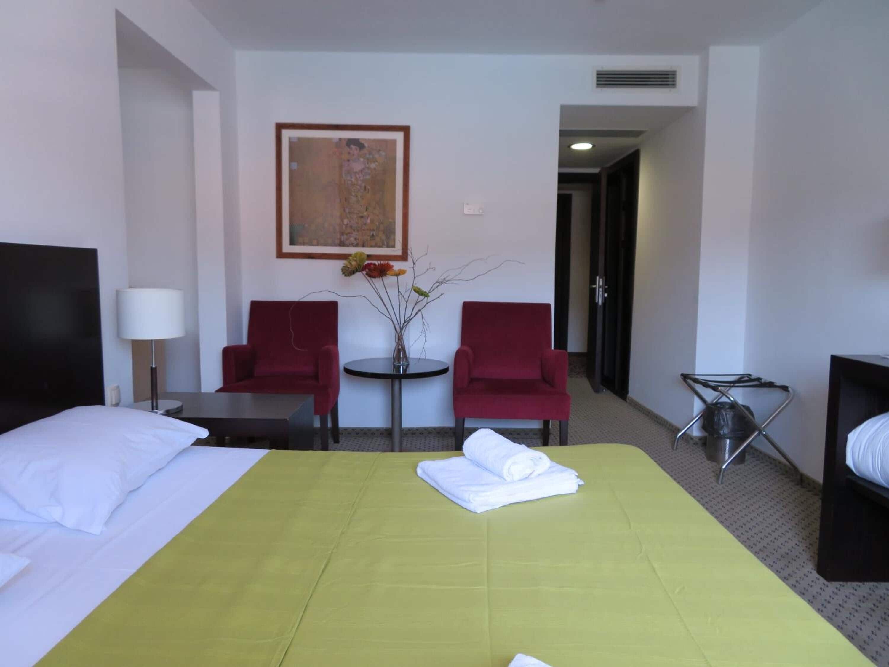 A hotel room with a bed, two red armchairs, a small table with a vase of flowers, and a painting on the wall.