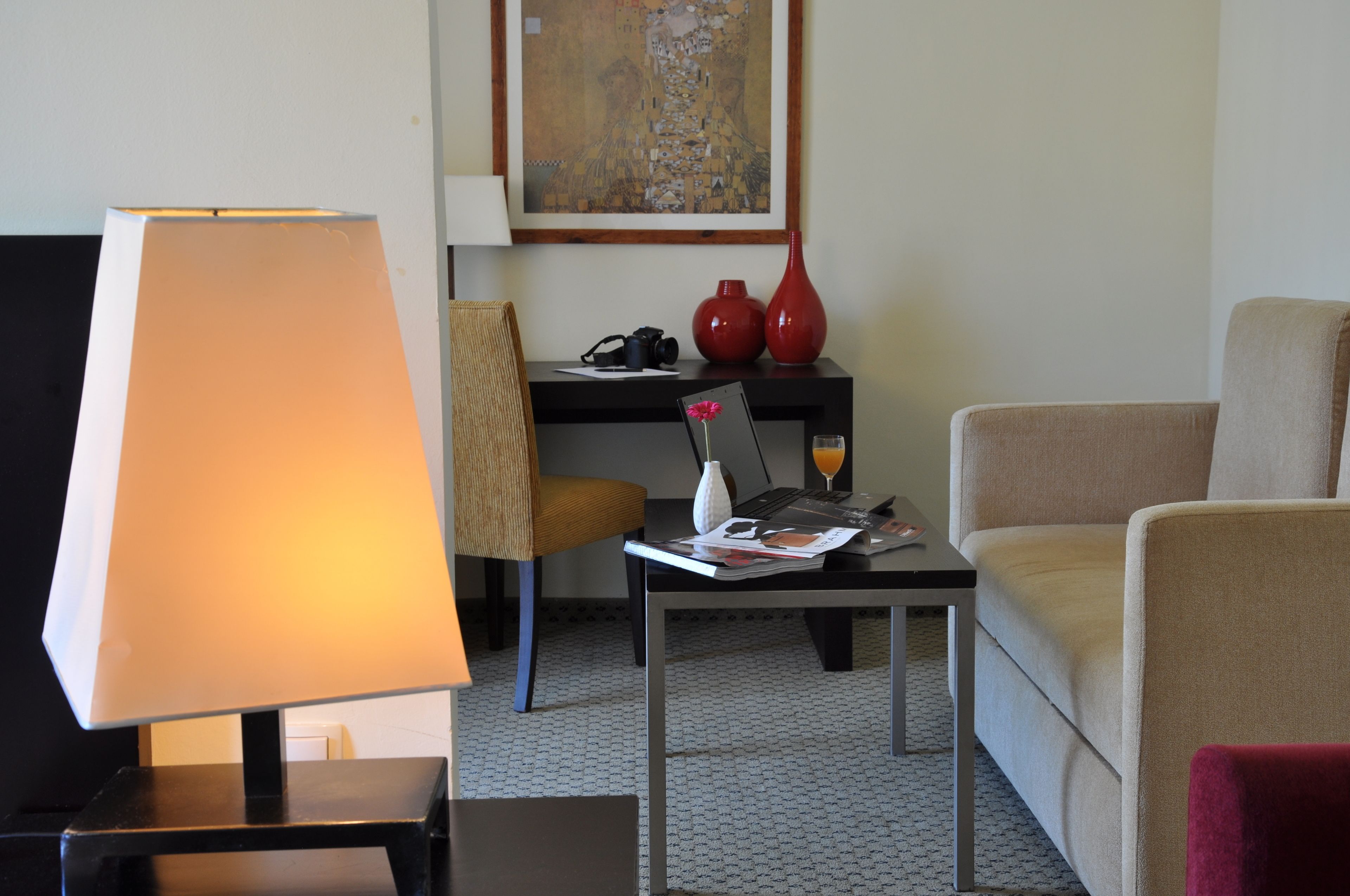 Hotellihuone, jossa on työpöytä, tuoli, sohva, lamppu, punainen maljakko ja taulu seinällä.
