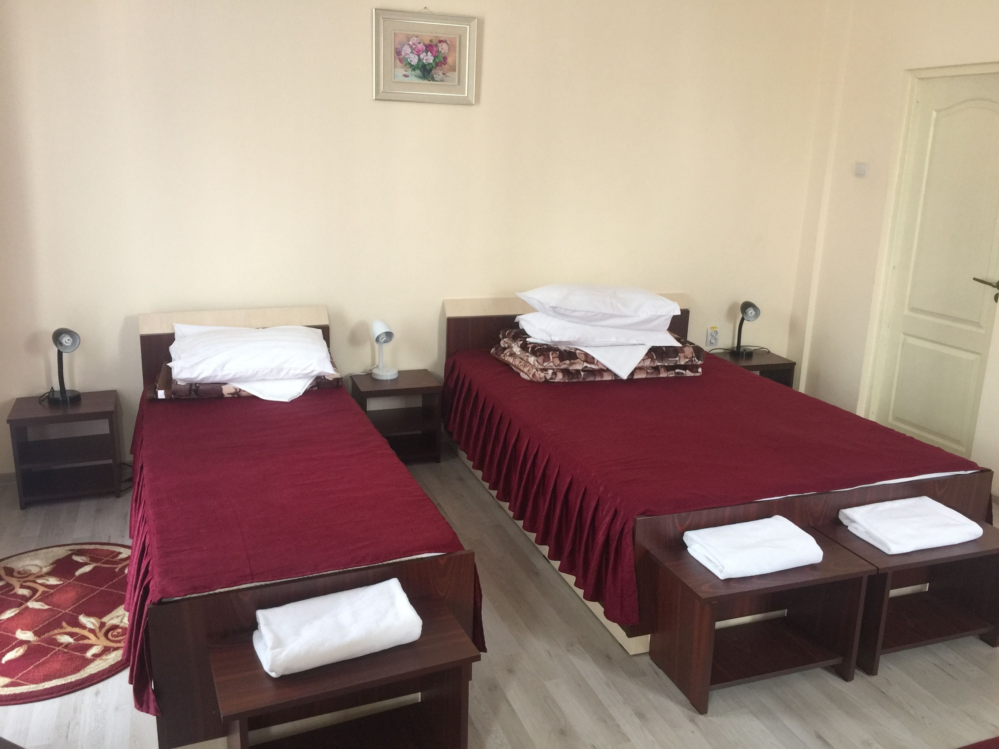 A hotel room with two single beds, each with a red bedspread and white pillows. There are bedside tables with lamps, a small rug, and a framed picture on the wall.