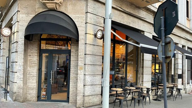 Un bâtiment en pierre avec un auvent noir et une entrée avec une porte en verre. Des tables et des chaises sont disposées à l’extérieur, sur le trottoir.