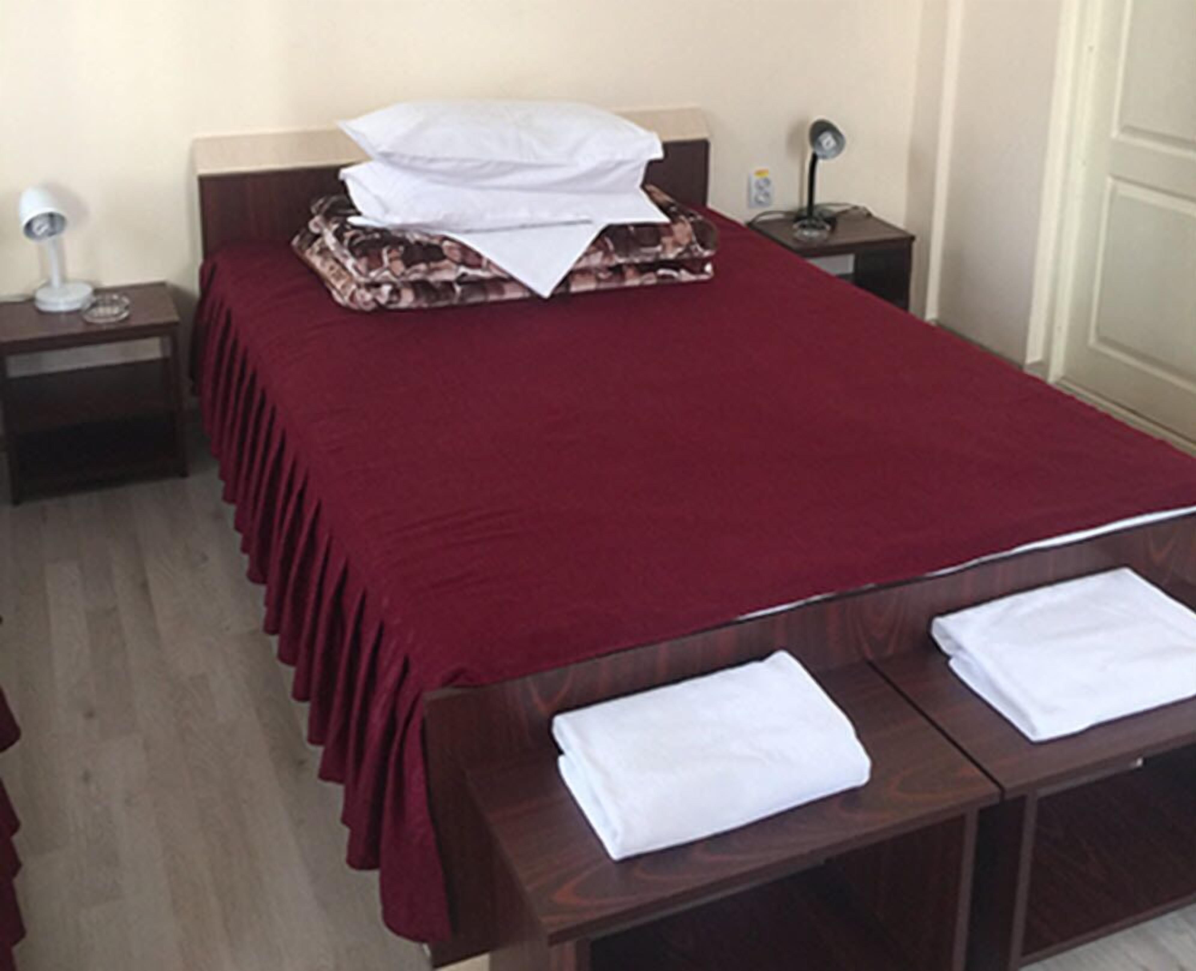 A neatly made bed with a red bedspread and white pillows, two bedside tables with lamps, and a wooden bench at the foot of the bed.