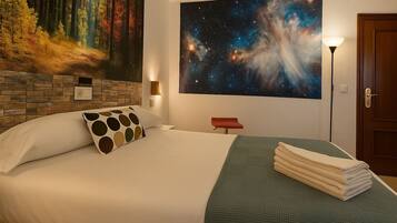 Una cama bien tendida con un almohadón de lunares, una manta verde con textura y una pila de toallas. Un cuadro grande de un bosque y una galaxia decora la pared.