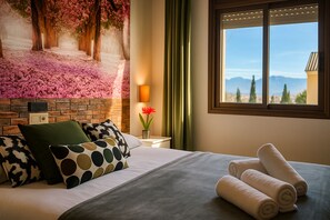 Habitación de hotel con cama, almohadas, mesita de noche con lámpara, ventana con vistas y un mural floral en la pared.