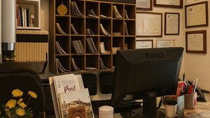Una zona de recepción con un mostrador, computadora, diplomas enmarcados y una estantería.