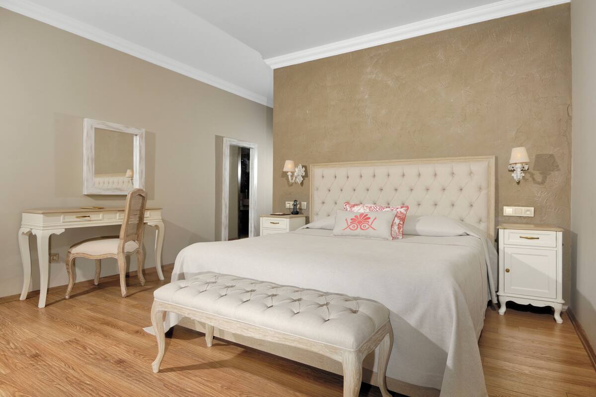 A bedroom with a large bed, a bedside table, a chair, a desk, and a mirror.