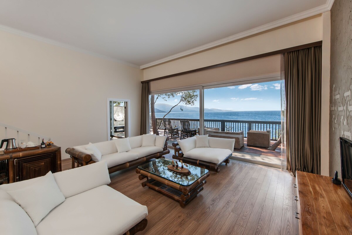 A spacious living room with a large sofa set, a coffee table, and a view of the sea through large windows.