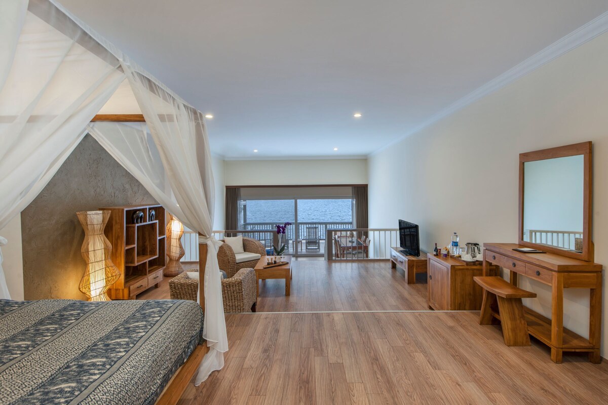 A bedroom with a bed, a wooden desk, a chair, a sofa, a balcony with a view, and a large window.