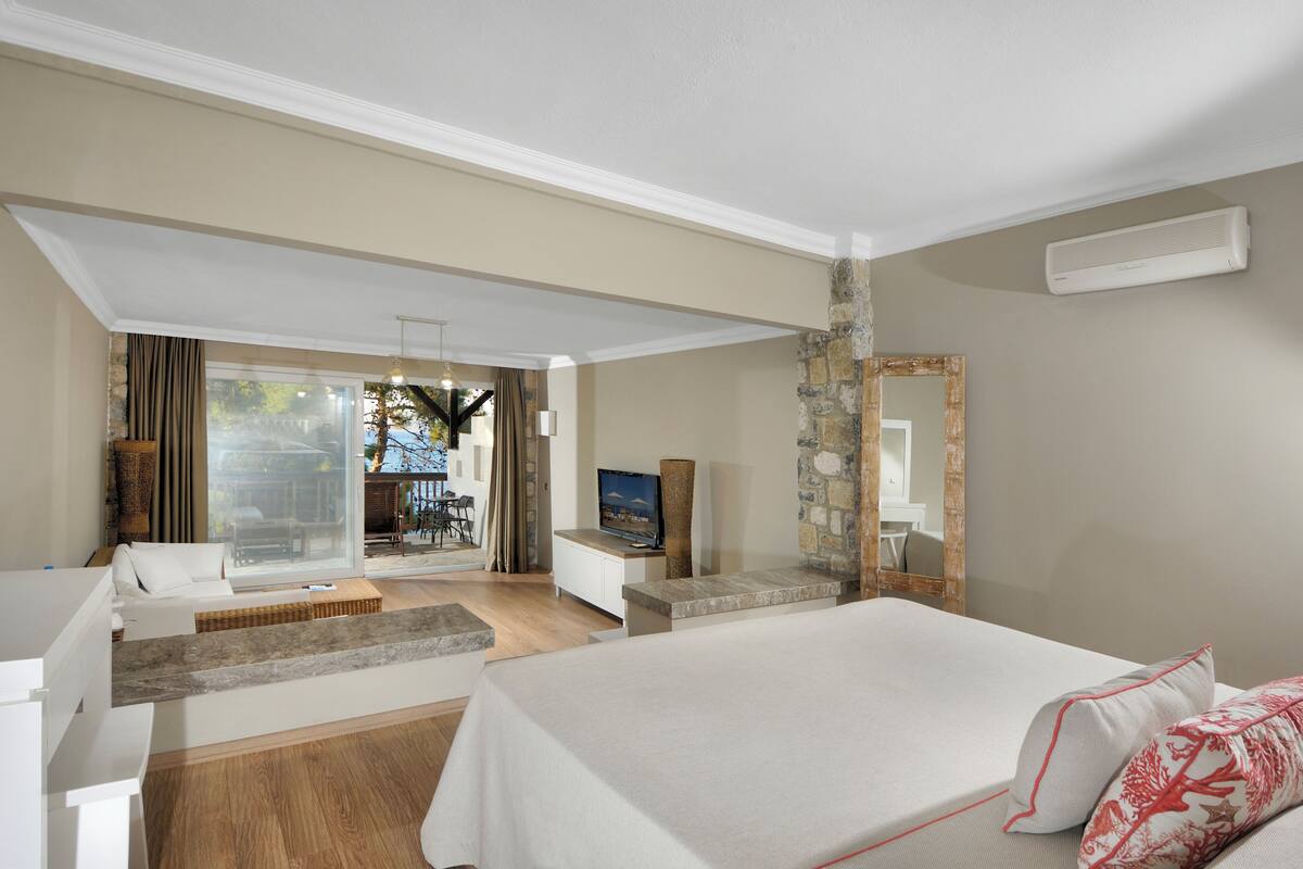 A modern hotel room with a bed, a TV, a stone accent wall, and a view of the outdoor area.