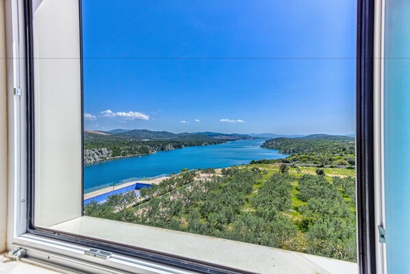 A view from a window overlooking a coastal landscape with a clear blue sky, a body of water, and a hilly terrain. - Hotel Panorama (Sibenik)