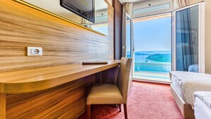A hotel room with a wooden desk, a chair, a bed, a TV, and a large window with a view of the sea. - Hotel Panorama (Sibenik)