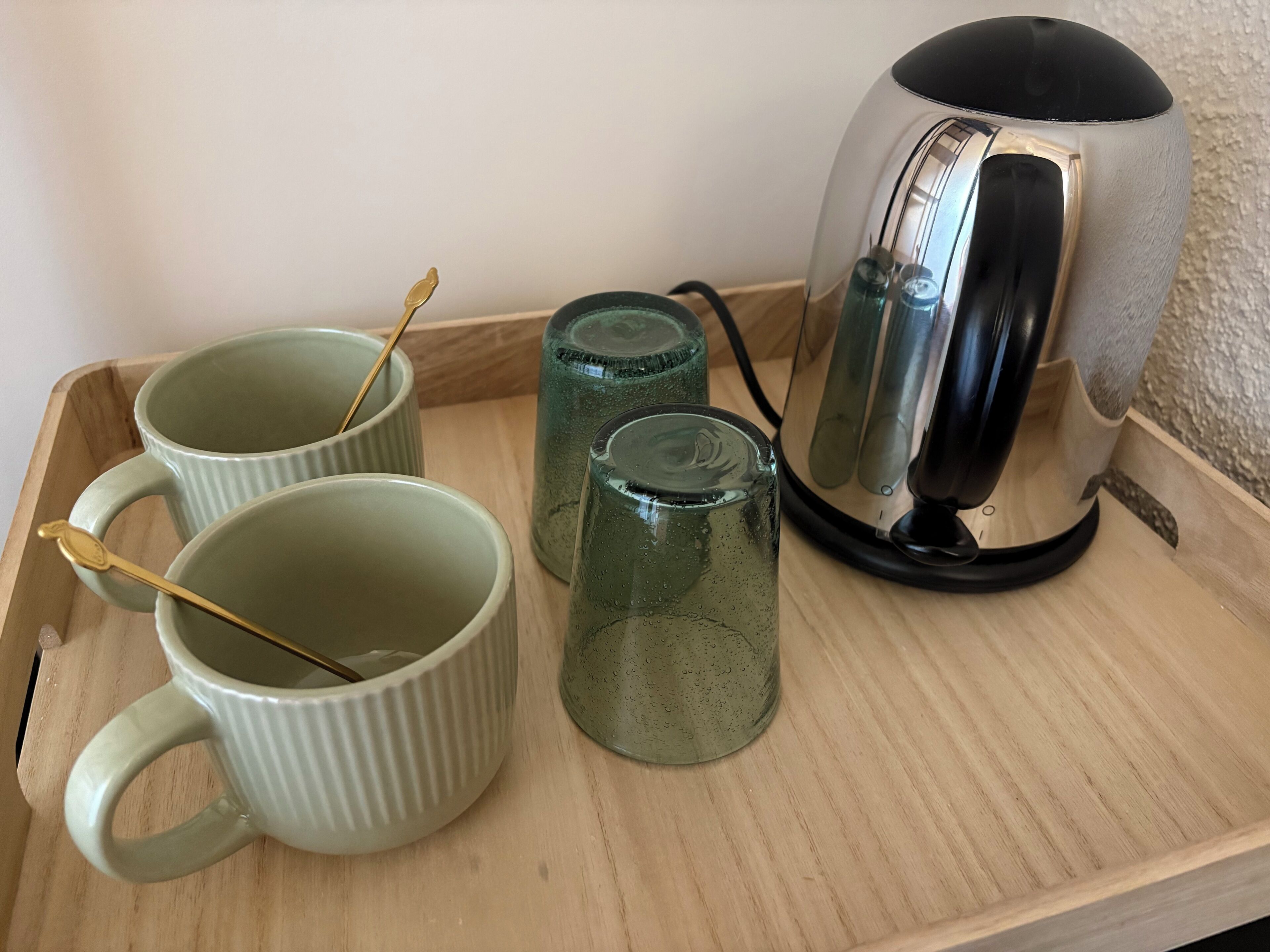 A tray with three ceramic mugs, two glass tumblers, and a kettle on a wooden surface.