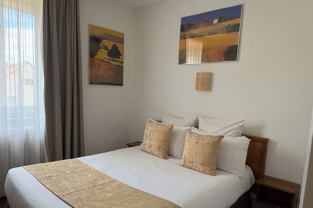 A hotel room with a large bed, two paintings on the wall, a bedside table, and a window with curtains.