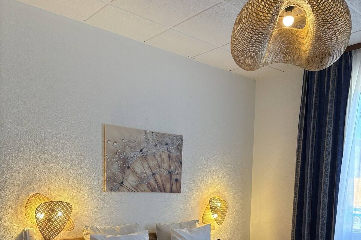 A hotel room with a large bed, two bedside tables, a wall-mounted lamp, and a window with curtains.