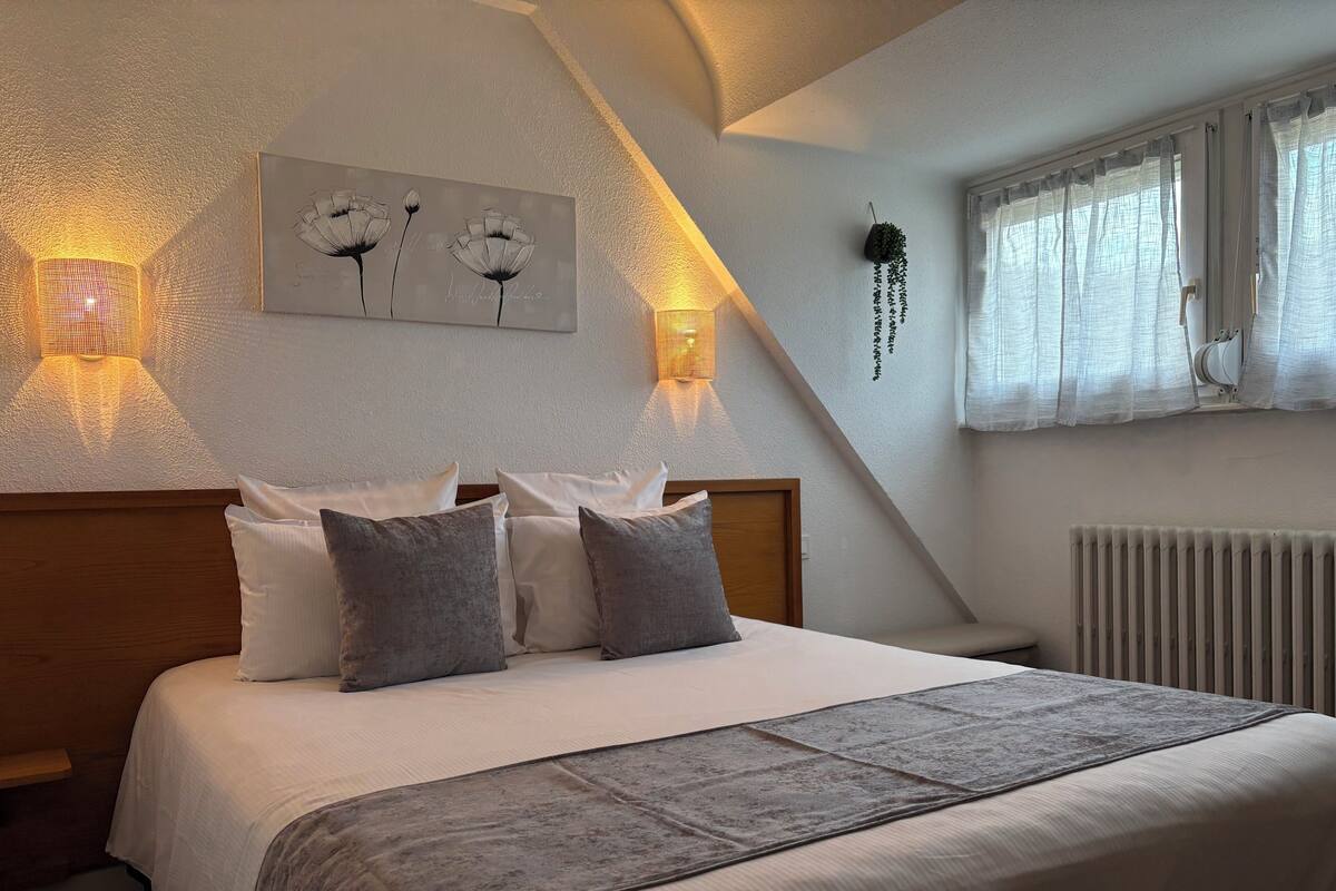 A bedroom with a bed, pillows, a bedside table, a wall-mounted light, a window with curtains, and a radiator.