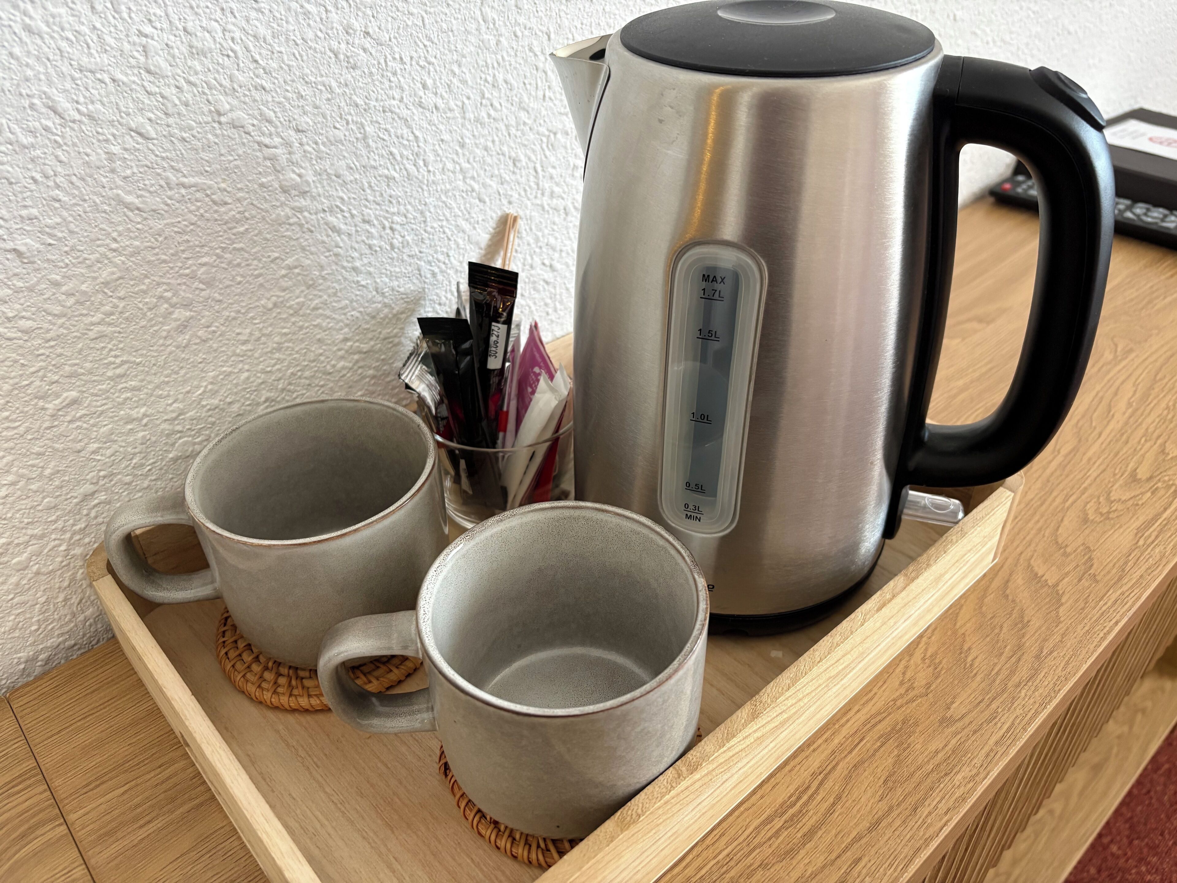 Ein Edelstahl-Wasserkocher, zwei graue Tassen und eine Schachtel mit Teebeuteln auf einem Holztisch.