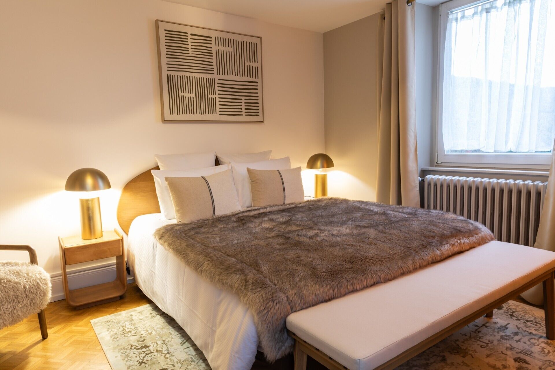 A bedroom with a bed, bedside lamps, a bench, a rug, and a window with curtains.
