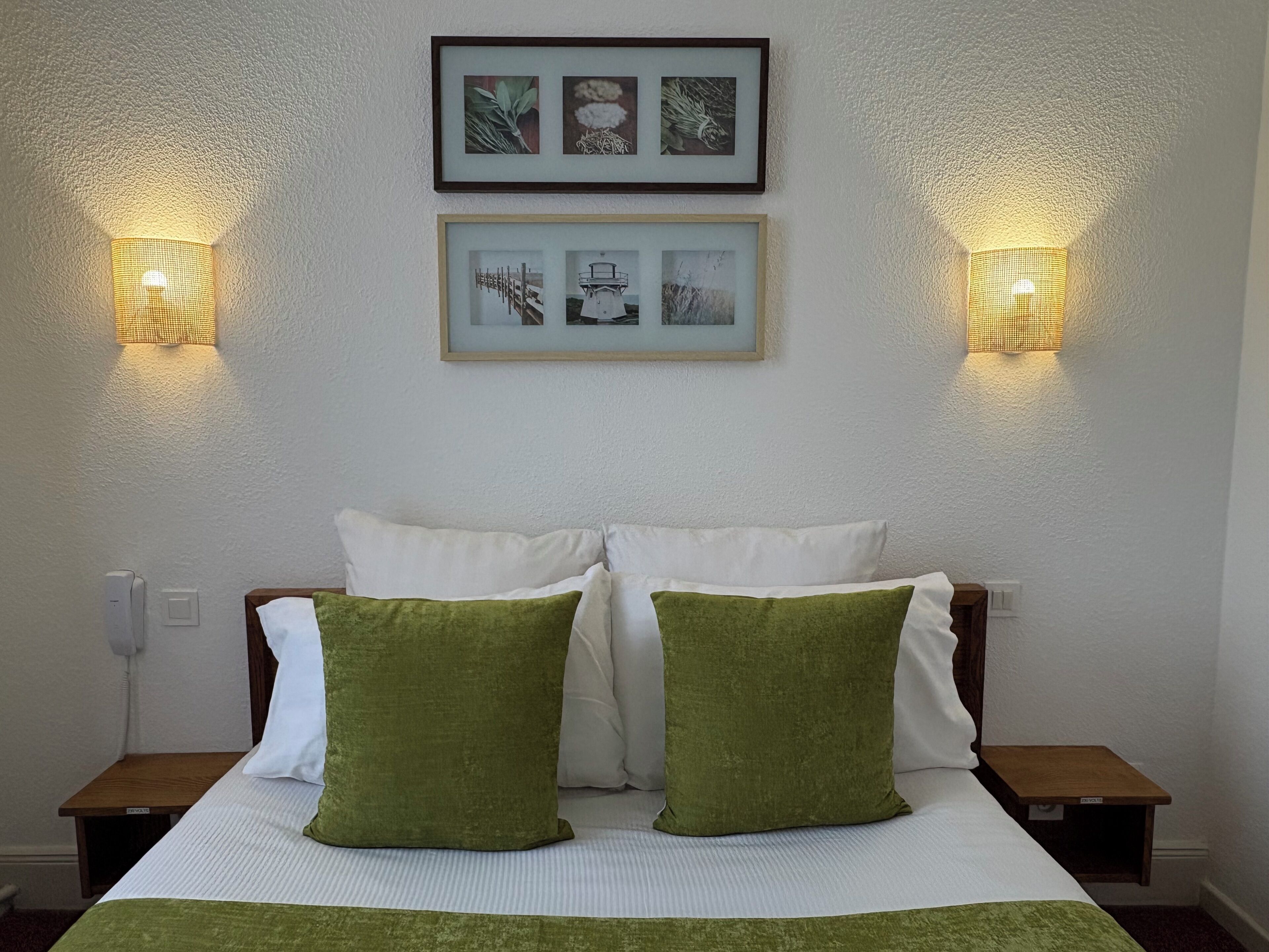 Une chambre d’hôtel comprenant un lit, deux tables de chevet, deux lampes fixées au mur et deux tableaux encadrés au-dessus du lit.